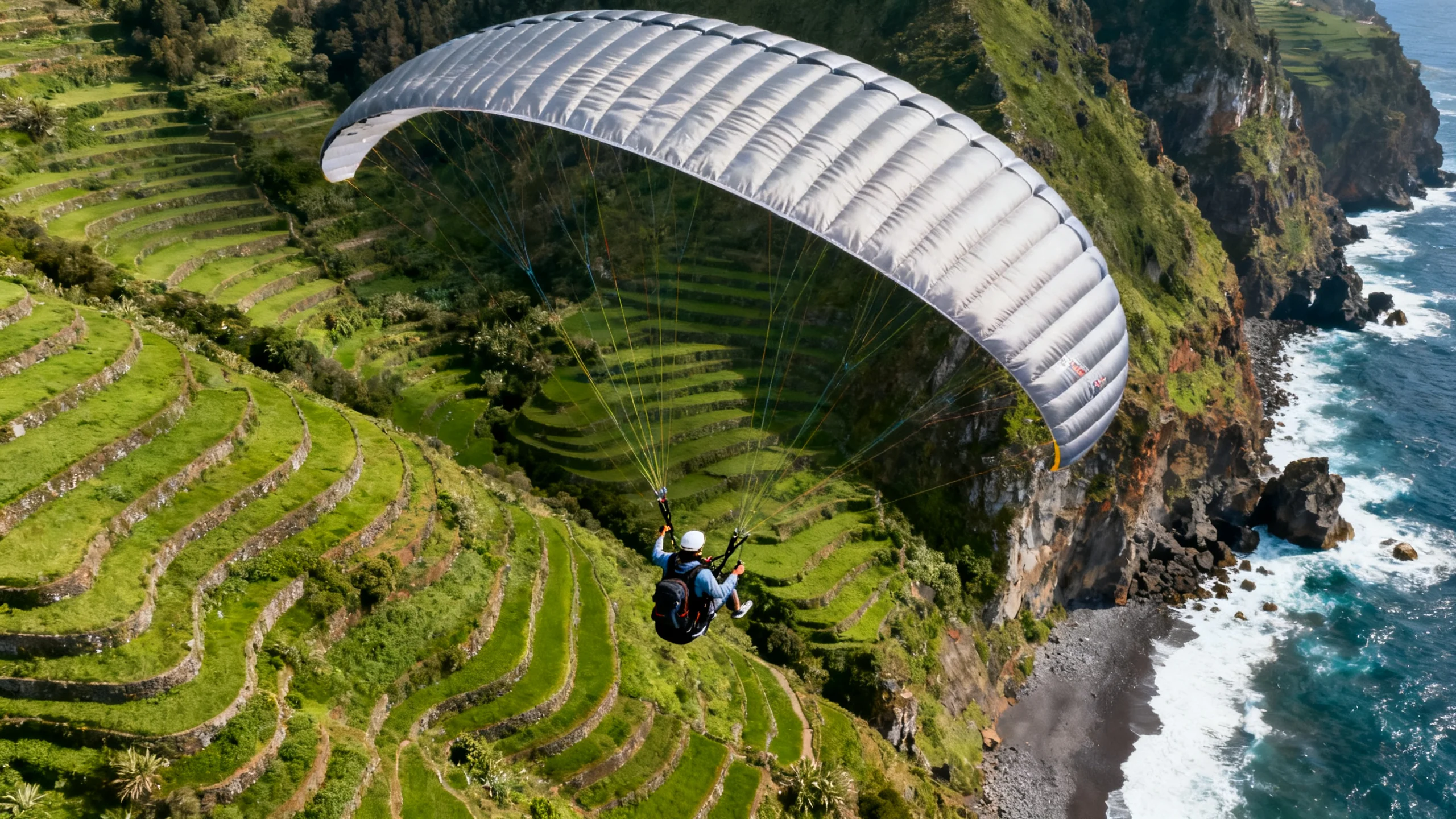 Un parapente biplaza en vuelo sobre las laderas verdes aterrazadas del oeste de Madeira con la costa atlántica debajo